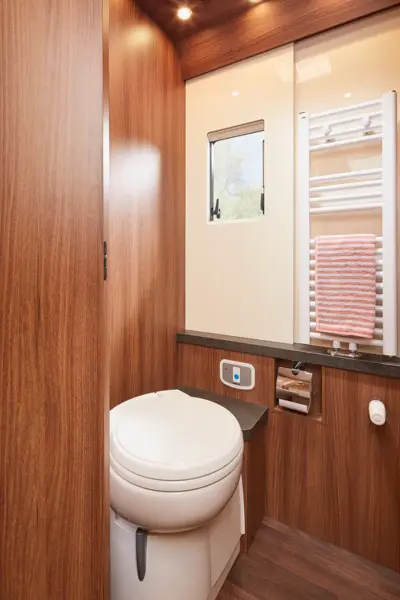 Modernes Wohnmobil-Badezimmer der Carthago Chic E Line mit Holzverkleidung, weißer Toilette, kleinem Fenster, wandmontiertem Handtuchheizkörper mit gestreiftem Handtuch, Toilettenpapierhalter und Bedienfeld. Die warme Beleuchtung schafft eine gemütliche Atmosphäre.