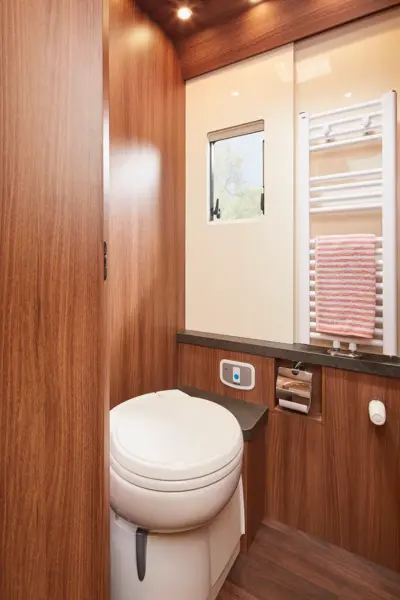 Ein kompaktes Badezimmer im Carthago Chic S Plus verfügt über holzvertäfelte Wände, eine weiße Toilette, ein kleines Fenster, einen Handtuchheizkörper mit einem gestreiften Handtuch und eingebaute Regale für Toilettenpapier und andere wichtige Dinge.