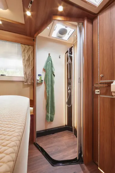Moderne Carthago Chic E Line Wohnmobil-Dusche mit hölzernem Interieur, einem grünen Handtuch an einem Haken, einer Regendusche und einem Bett mit gestepptem beigen Bezug in der Nähe. Ein Fenster mit Vorhängen bringt natürliches Licht herein.