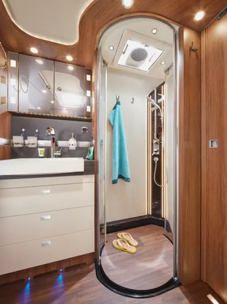 Modernes Wohnmobil-Badezimmer im Carthago Chic E Line mit einer gebogenen Glasdusche, Holzwänden, einem weißen Waschbecken mit Schränken, Deckenleuchten, einem blauen Handtuch in der Dusche und einem Paar Sandalen auf dem Holzboden.