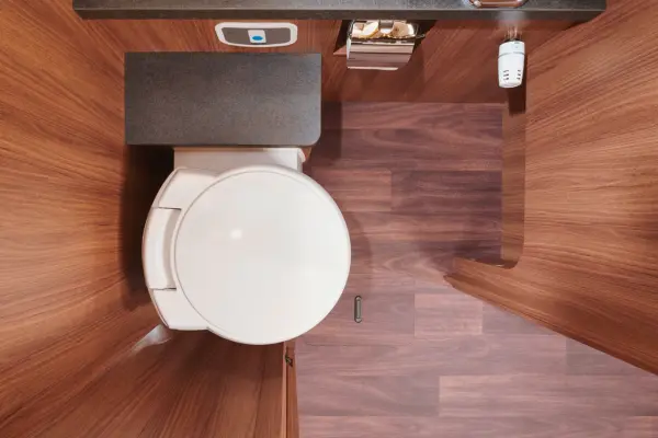 Draufsicht auf ein kleines, modernes Badezimmer mit holzgetäfelten Wänden und Böden in einem Carthago Chic E Line, mit weißer Toilette, grauer Arbeitsplatte, Toilettenpapierhalter mit Papier und einem kleinen Lufterfrischer auf dem Tresen.