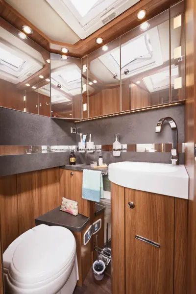 Modernes Carthago Chic S Plus Wohnmobil-Badezimmer mit Holzschränken, einem weißen Waschbecken, einer kompakten Toilette, großen Spiegeln, Handtuchhaltern und Oberlichtern für natürliches Licht. Toilettenartikel und Handtücher sind übersichtlich angeordnet.