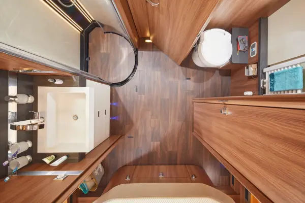 Draufsicht auf ein kompaktes, modernes Carthago Chic S Plus Wohnmobil-Badezimmer mit Holzverkleidung, einem quadratischen weißen Waschbecken, einer Glastür in der Dusche, einer Toilette, Toilettenartikeln, Ablagen und einem Handtuchhalter mit einem blauen Handtuch.