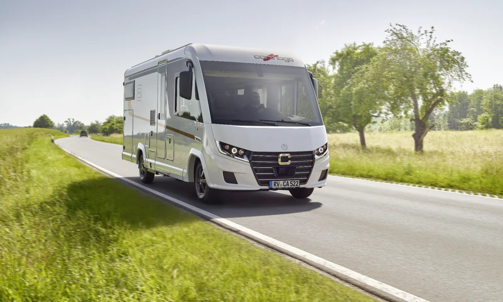 Ein weißes vollintegriertes Wohnmobil fährt auf einer zweispurigen Landstraße, gesäumt von grünem Gras und Bäumen, unter einem klaren, sonnigen Himmel.