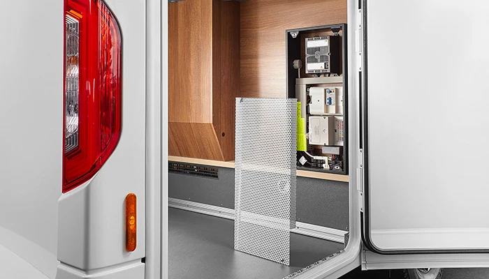 Innenansicht eines Fahrzeugstauraums mit Holzverkleidung, einer Metallgitterabdeckung und verschiedenen elektronischen Bauteilen und Kabeln, die hinter einer offenen Zugangsplatte sichtbar sind.