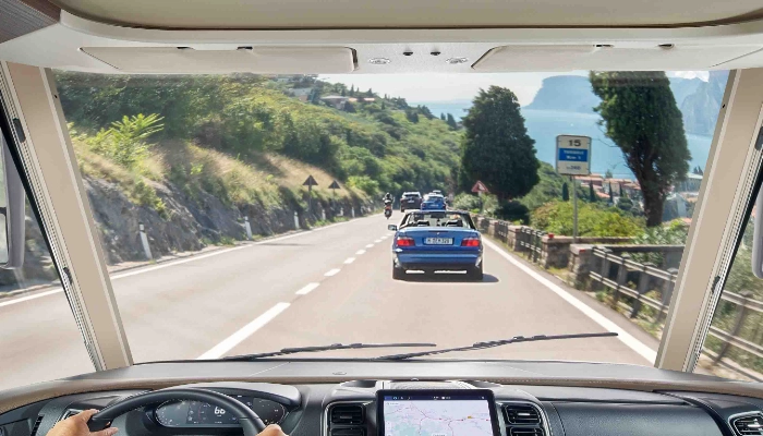 Vista dall’interno di un veicolo che percorre una strada panoramica e tortuosa, con altre automobili davanti, verde e alberi su entrambi i lati e uno specchio d’acqua e montagne sullo sfondo.
