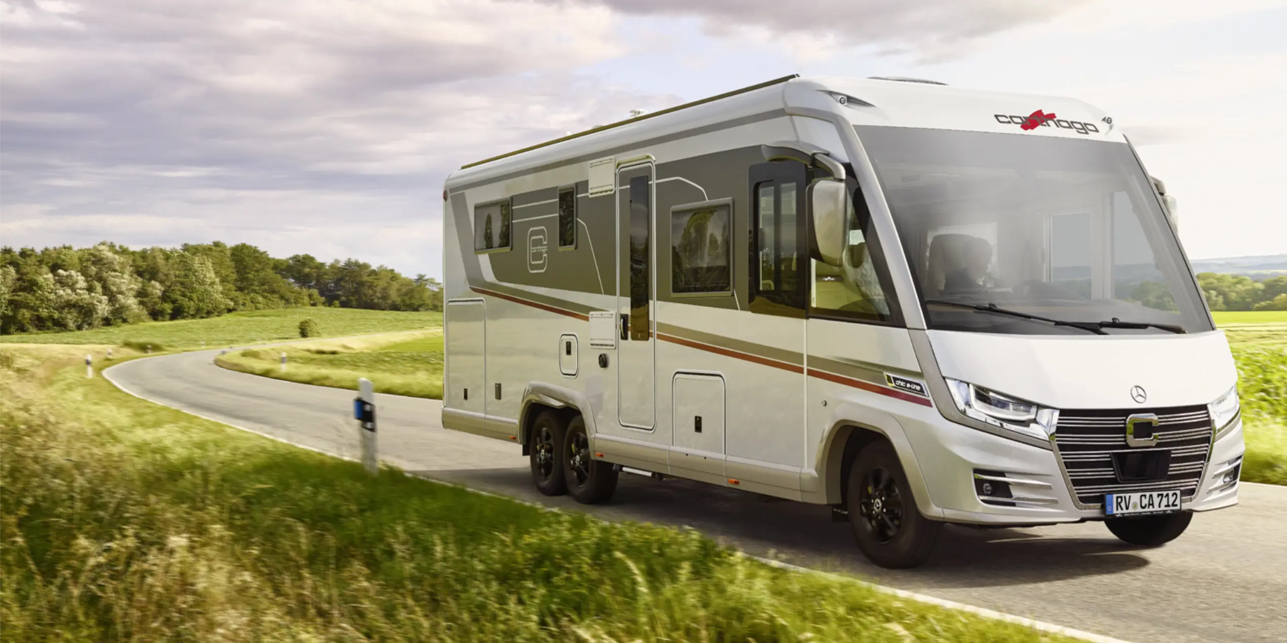 Ein modernes, weißes vollintegriertes Wohnmobil fährt auf einer kurvigen Landstraße durch eine grüne Landschaft mit Bäumen und Feldern unter einem teils bewölkten Himmel.