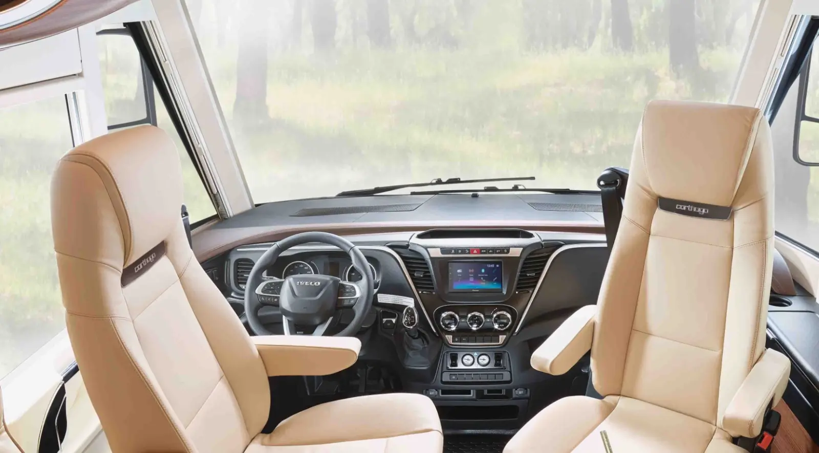 The inside of a motorhome cab with two beige Captain seats, a modern dashboard with digital display, steering wheel and large windows, showing a blurry, forest-like background.