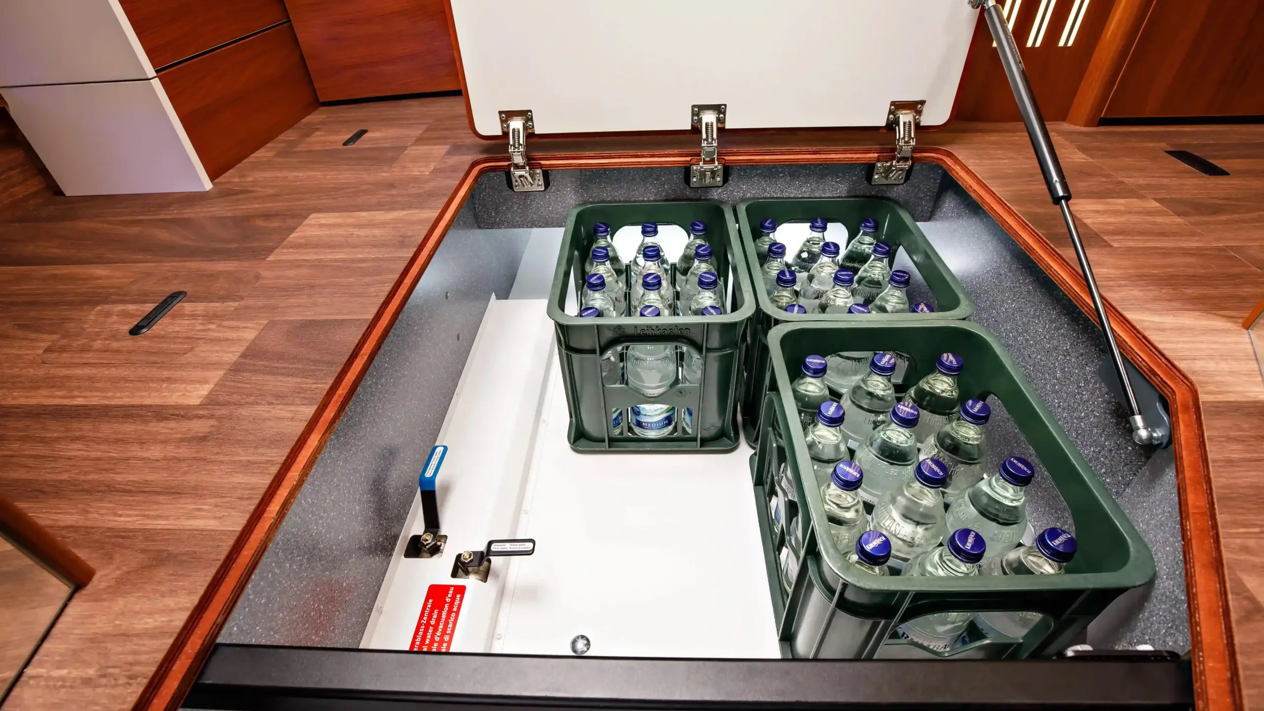 Ein offenes Staufach im Holzboden enthält drei grüne Kisten mit blauen Wasserflaschen, die sich nahtlos in das elegante Interieur eines Carthago Liner for Two einfügen.