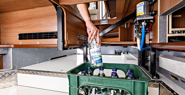 Die Hand einer Person stellt eine Glasflasche mit Mineralwasser in eine grüne Kiste unter einem hölzernen Staufach in einem Wohnmobil, in dem verschiedene Rohre und Geräte sichtbar sind.