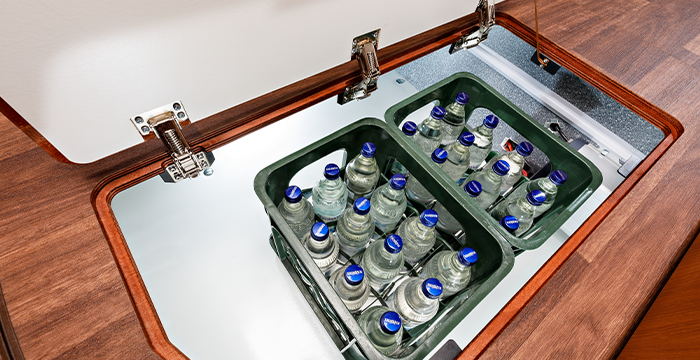 Zwei mit Glasflaschen gefüllte Kisten mit Wasser werden in einem eingelassenen Fach mit einer Holzplatte und Metallscharnieren aufbewahrt, das in eine hölzerne Arbeitsplatte eingebaut ist.