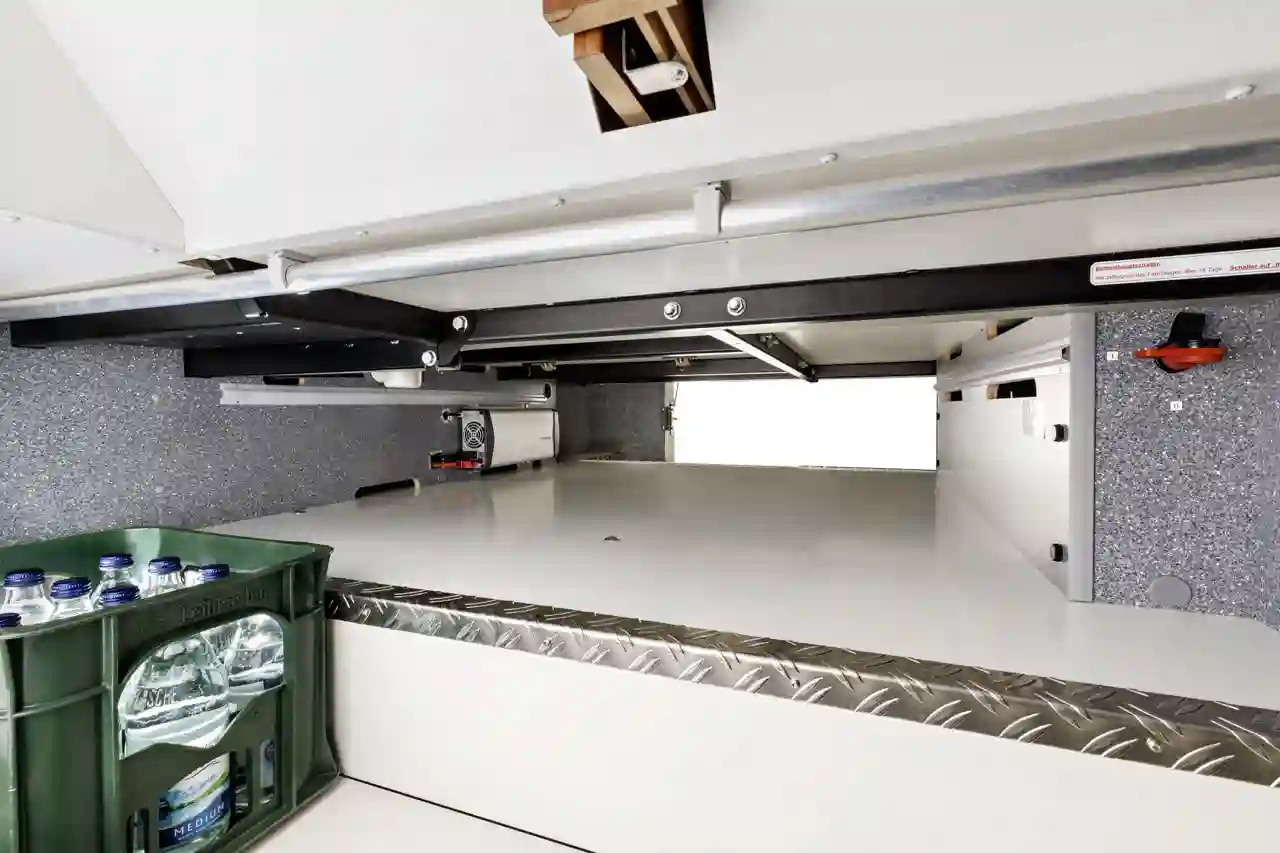 Innenansicht eines Carthago Liner for Two Stauraums mit einer grünen Kiste mit Wasserflaschen auf der linken Seite, Metallverkleidungen am Boden und sichtbaren Regalen, Schienen und Kabeln an den Seiten und an der Decke.