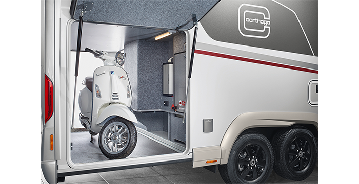 Uno scooter bianco è parcheggiato nel vano di stivaggio di un moderno rimorchio da viaggio Carthago Chic S Plus bianco e grigio con ruote nere e un logo Carthago sul fianco.