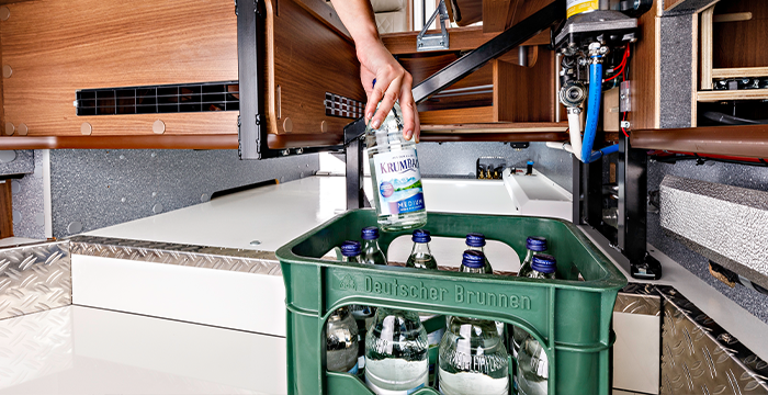 Una persona mette una bottiglia di vetro contenente acqua minerale in una cassa verde, riempita con bottiglie simili, in un moderno camper Carthago Chic S Plus con armadi in legno e vani di stivaggio visibili.