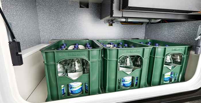 Drei grüne Plastikkisten, die jeweils mehrere Glasflaschen mit Wasser enthalten, sind ordentlich in einem Fach mit metallischer Innenausstattung des teilintegrierten Wohnmobils untergebracht.