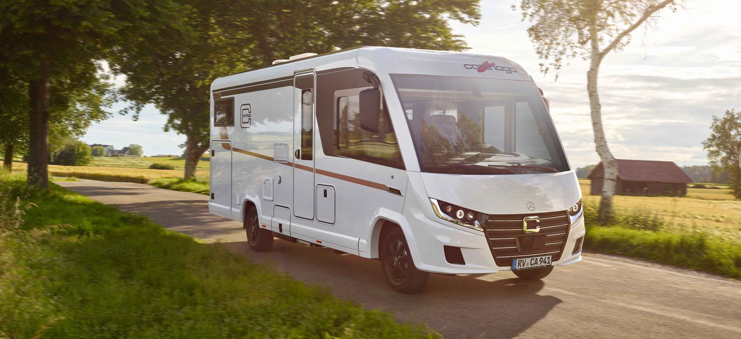 Ein weißes vollintegriertes Wohnmobil fährt entlang einer malerischen Landstraße, die von Bäumen und grasbewachsenen Feldern gesäumt ist, unter einem teilweise bewölkten Himmel.