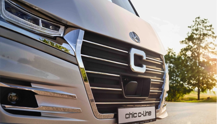 Nahaufnahme des Kühlergrills eines modernen integrierten Wohnmobils mit einem großen stilisierten C in der Mitte und "chic c-line" auf dem Nummernschild, Bäume und Sonnenlicht im Hintergrund.