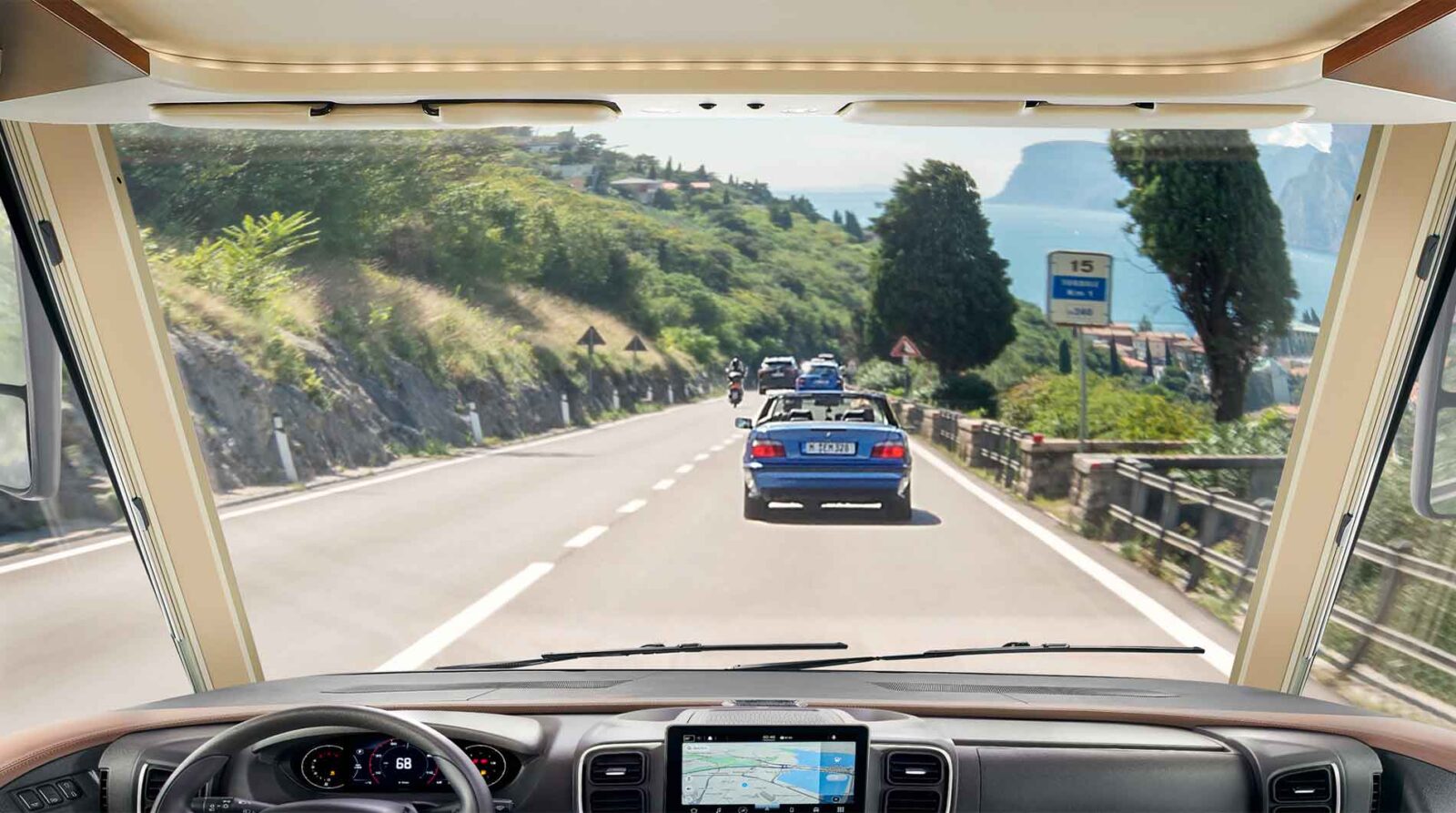 Blick aus dem Inneren eines vollintegrierten Wohnmobils, das eine kurvenreiche, von Bäumen und Leitplanken gesäumte Küstenstraße entlangfährt und mehreren Autos folgt, mit malerischen Hügeln und Wasser im Hintergrund. Ein GPS-Navigationsbildschirm befindet sich auf dem Armaturenbrett.