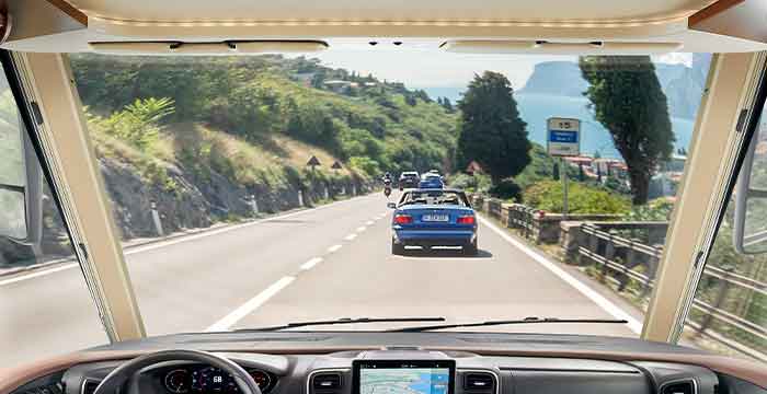 Blick aus dem Inneren eines integrierten Wohnmobils, das auf einer malerischen, kurvenreichen Straße mit vorausfahrenden Autos fährt, umgeben von Grünflächen, Hügeln und fernen Bergen unter einem klaren Himmel. Ein Navigationsbildschirm ist auf dem Armaturenbrett zu sehen.