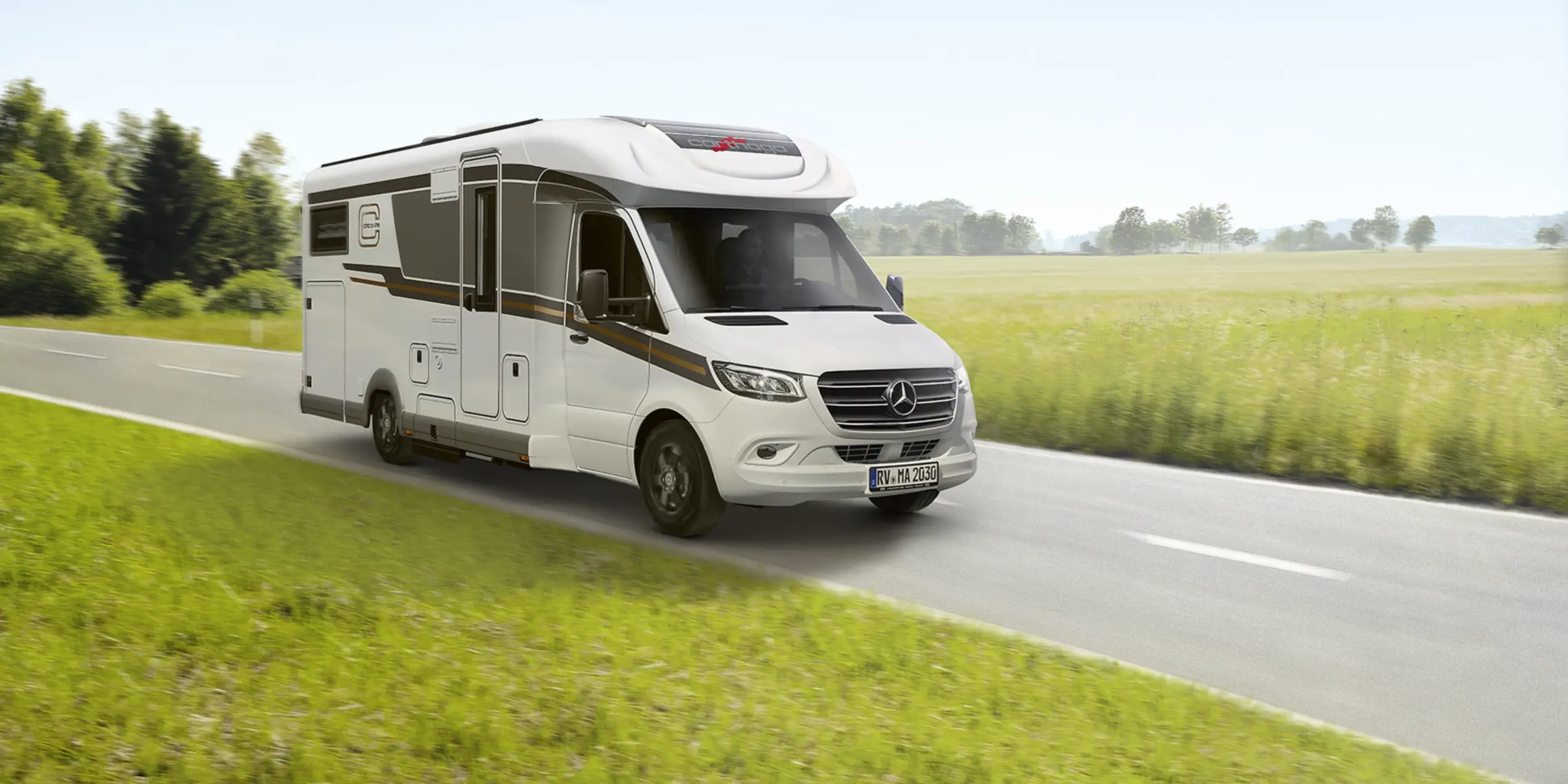 Ein weißes Wohnmobil fährt eine Landstraße entlang, die von grünem Gras und Feldern gesäumt ist, mit Bäumen und einem blauen Himmel im Hintergrund.