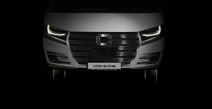 Frontansicht eines modernen vollintegrierten Wohnmobils mit beleuchteten LED-Scheinwerfern, einem großen schwarzen Kühlergrill und einem Nummernschild mit der Aufschrift chic e-line vor einem dunklen Hintergrund.