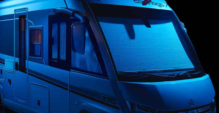 Vorderansicht der Fahrerseite eines integrierten Wohnmobils, das in kühles, tiefblaues Licht getaucht ist. Der Fokus liegt auf der geschlossenen Frontscheiben-Verdunkelung, die vollständig heruntergezogen ist und die gesamte Windschutzscheibe bedeckt. Auch das Seitenfenster auf der Fahrerseite ist durch ein geschlossenes Plisseerollo vor Einblicken geschützt. Man sieht die Aufbautür mit Fenster und die markanten, hängenden Busspiegel. Das Logo des Herstellers ist über der Frontscheibe erkennbar