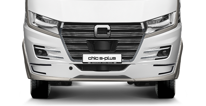 Vista frontale di un camper di lusso Carthago Chic S Plus bianco con eleganti fari, una griglia del radiatore nera pronunciata e una targa con la scritta Chic S-Plus. Il design del veicolo si distingue per le sue linee moderne e spigolose.