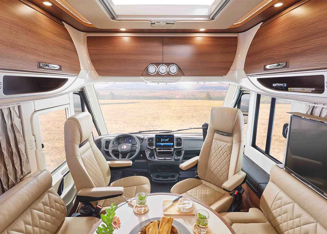 Der Innenraum dieses integrierten Wohnmobils besteht aus beigen Ledersitzen, einem Tisch mit Lebensmitteln und Blumen, Holzschränken, einem großen Fenster mit Blick auf ein Feld und einem eleganten Fahrerplatz.