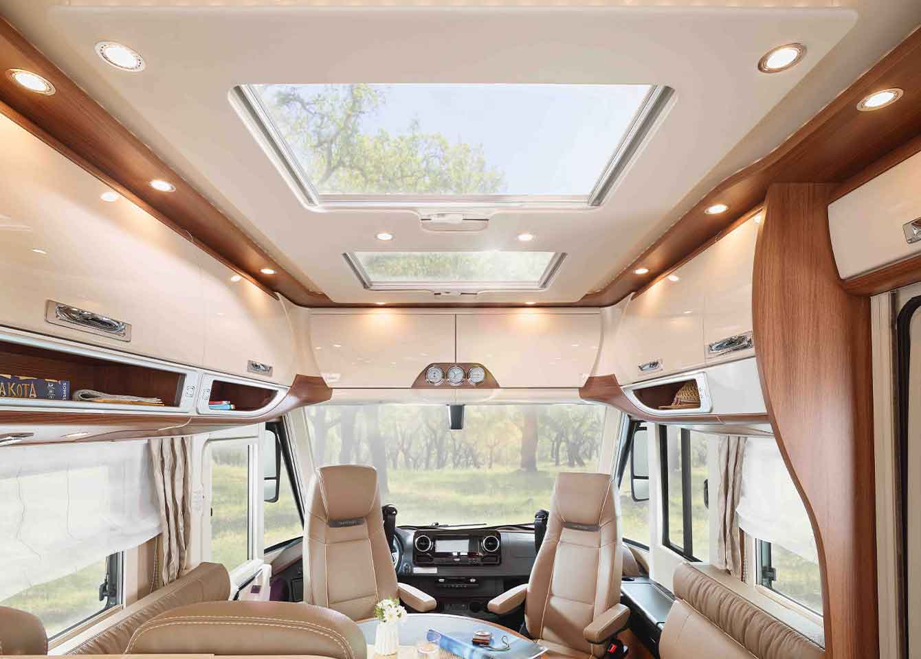 Das Innere dieses luxuriösen teilintegrierten Wohnmobils ist mit Ledersitzen, großen Fenstern, Holzakzenten, Oberlichtern und Deckenleuchten ausgestattet, die eine geräumige und helle Atmosphäre mit einem schönen Blick auf die Bäume im Freien schaffen.