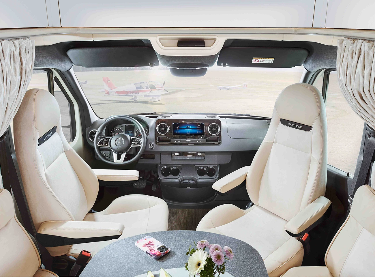 Innenansicht eines modernen Wohnmobils mit zwei cremefarbenen Sitzen, die einem kleinen runden Tisch gegenüberstehen, einem Armaturenbrett mit Lenkrad und einem Fenster, aus dem Flugzeuge und eine Startbahn zu sehen sind. Auf dem Tisch stehen eine Vase mit Blumen und ein Telefon.