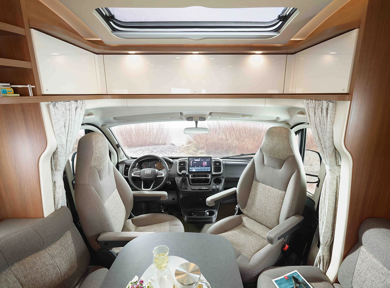 Innenraum eines modernen Wohnmobils mit Fahrer- und Beifahrersitz, einem zentralen Tisch mit Glas und Schüssel, Stauschränken und einem großen Fenster darüber, das für Tageslicht sorgt.