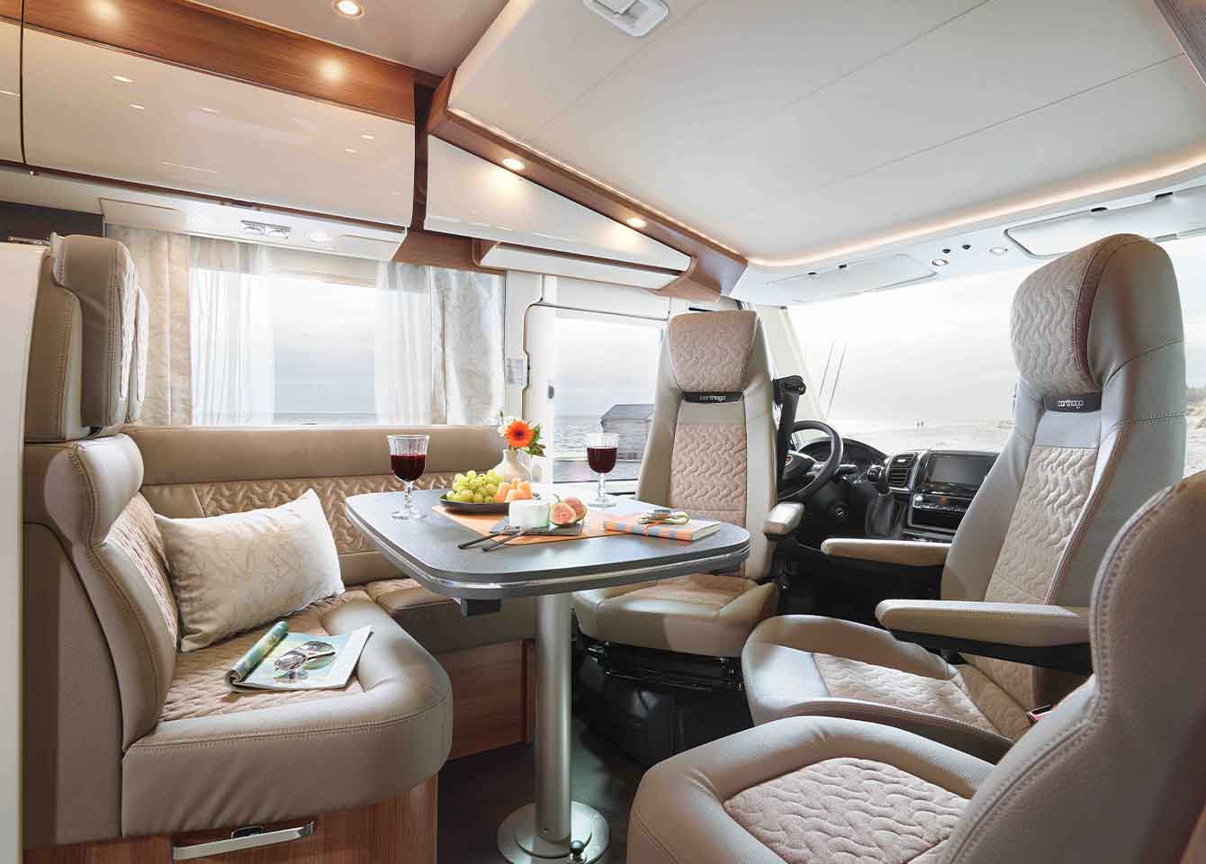 Geräumiges, vollintegriertes Wohnmobil-Interieur mit einem Tisch für mehrere Personen, an dem Wein, Obst und Blumen stehen. Plüschige graufarbene Sitze umgeben den Tisch, große Fenster lassen natürliches Licht herein. Auf einem der Sitze liegt eine Zeitschrift.