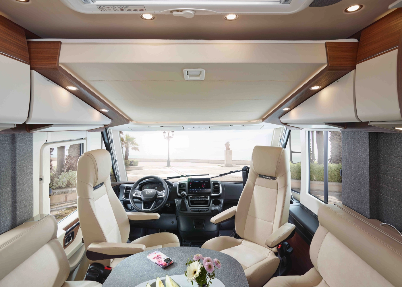 Innenraum eines modernen Wohnmobils mit cremefarbenen Sitzen, einem runden Tisch mit Blumen und Snacks, großen Fenstern und Blick auf Palmen und die Strandpromenade draußen.