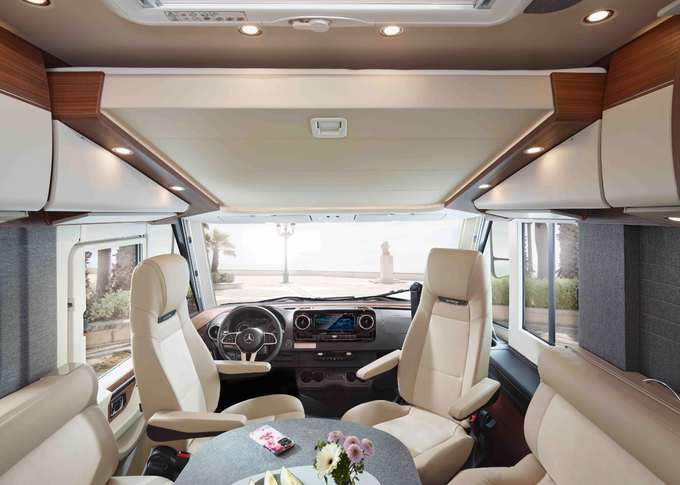 Modernes Wohnmobil-Interieur mit vier cremefarbenen Drehsesseln um einen kleinen runden Tisch, einer großen Frontscheibe, die eine malerische Aussicht auf das Meer mit Palmen bietet, und Staufächern im Dach.