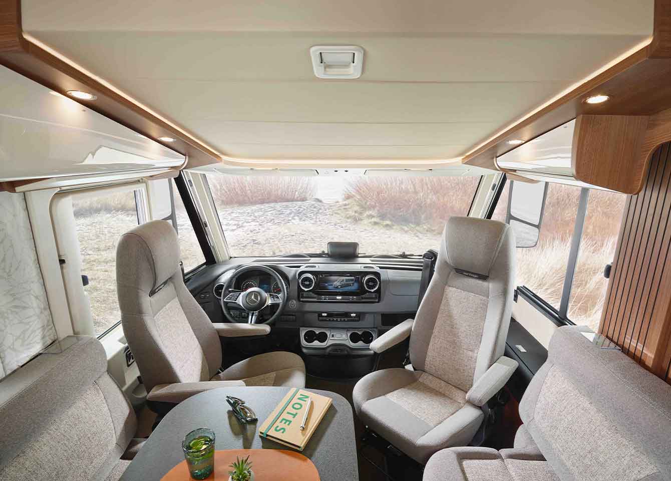 Der Innenraum eines modernen vollintegrierten Wohnmobils mit Fahrer- und Beifahrersitz, einem Tisch mit Notebook und Getränken sowie großen Fenstern, die den Blick auf Gras und Wasser freigeben.