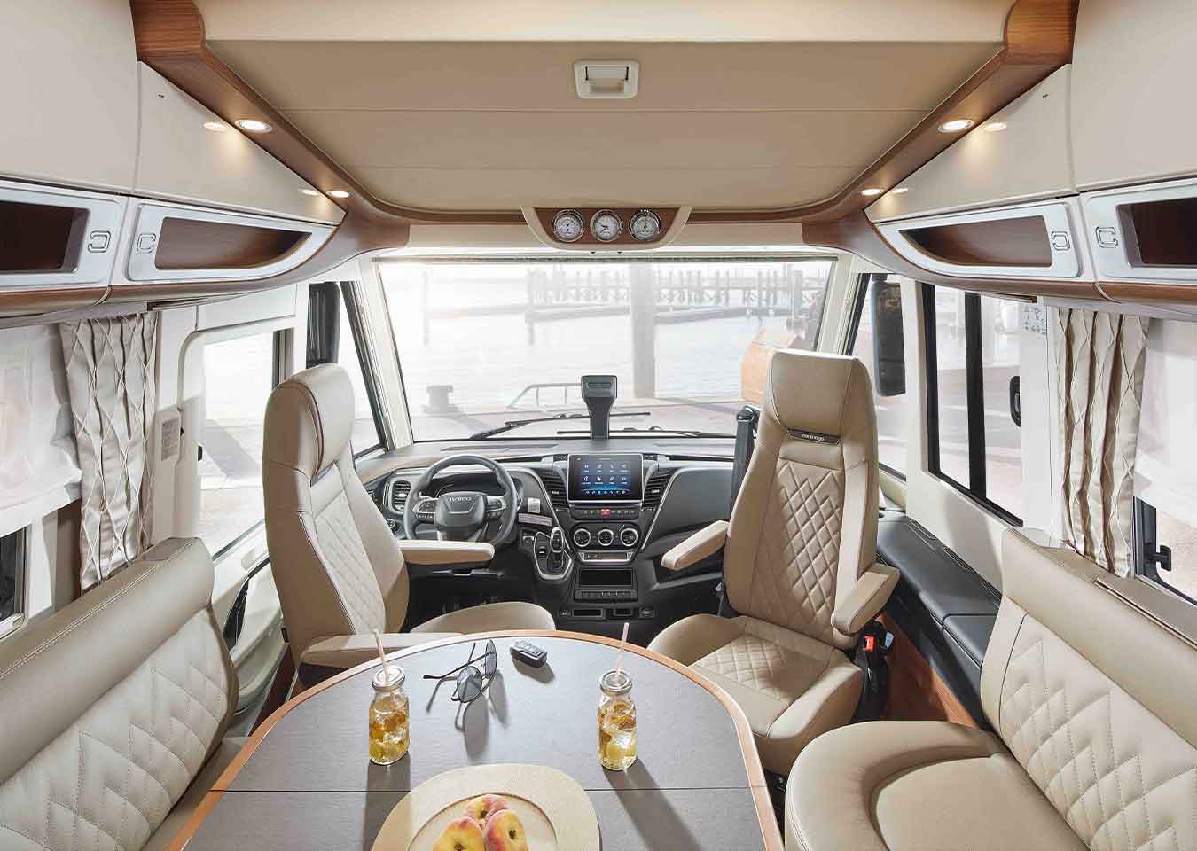 Innenraum eines modernen Wohnmobils mit beigen Ledersitzen, einem Esstisch mit Obst und Getränken, großen vorderen Fenstern mit Vorhängen und Blick auf das Wasser draußen. Das Cockpit des Fahrers und das Armaturenbrett sind sichtbar.