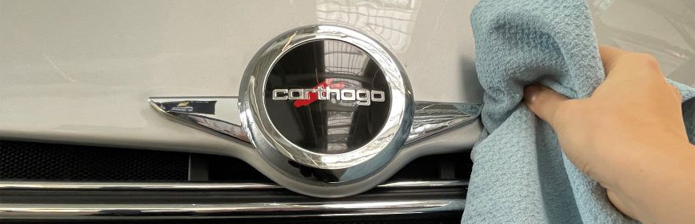 Eine Hand poliert mit einem hellblauen Tuch das vordere Emblem eines silbernen Autos mit der Aufschrift Carthago und sorgt dafür, dass es genauso glänzt, wie wenn man über das Carthago Hilfe Portal Hilfe sucht.