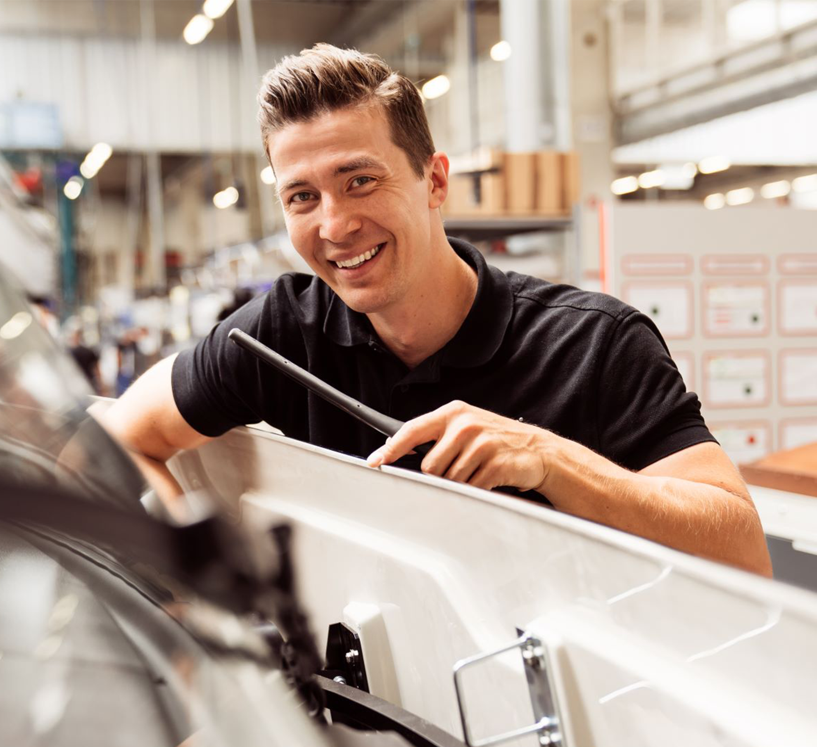 Ein lächelnder Mann in einem schwarzen Polohemd arbeitet an der Montage oder Reparatur von Maschinen in einer hell erleuchteten Industriehalle. Er hält ein Werkzeug in der Hand und steht neben einer großen weißen Maschine. Möglicherweise verweist er auf Anweisungen aus dem Carthago Hilfe Portal.