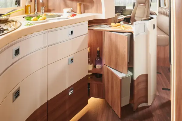 Eine moderne Wohnmobilküche in einem Carthago Chic E Line verfügt über geschwungene Holzschränke, offene Schubladen mit Flaschen und Stauraum, ein Schneidebrett mit Brot und Gemüse und einen eingebauten Kühlschrank. Der Raum wirkt sauber, hell und aufgeräumt.