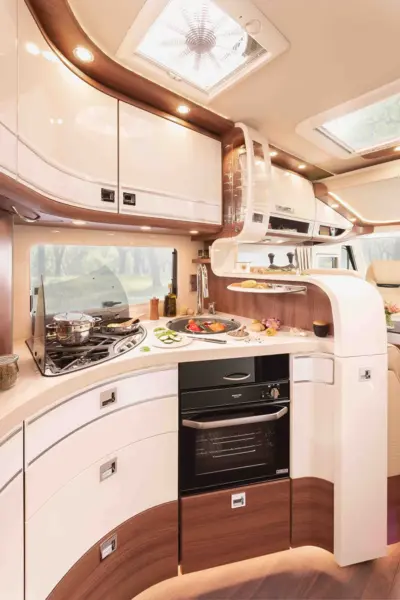 Moderne Wohnmobilküche der Carthago Chic E Line mit glänzend weißen Schränken, einem Gasherd mit Töpfen, Backofen und Geschirr auf einer geschwungenen Arbeitsplatte. Das Sonnenlicht fällt durch die Oberlichter auf das frische Gemüse. Durch das Fenster sind Bäume zu sehen.