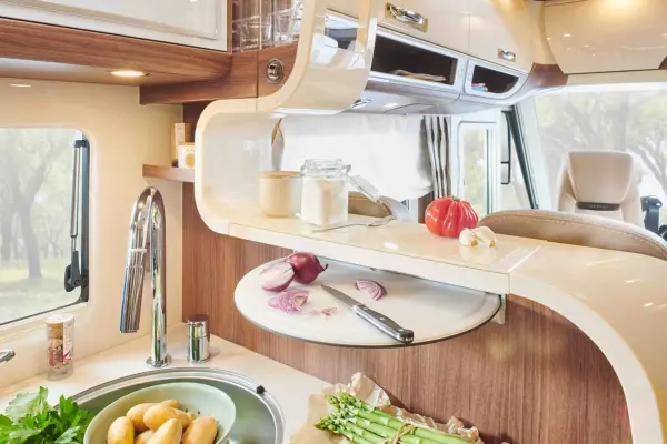 Eine moderne Wohnmobilküche im Carthago Chic E Line verfügt über hölzerne und weiße Schränke, ein rundes ausziehbares Schneidebrett mit gehackten roten Zwiebeln, Knoblauch und Tomaten. Frisches Gemüse wie Kartoffeln und Spargel stehen neben der Spüle und einer Dose mit Mehl.