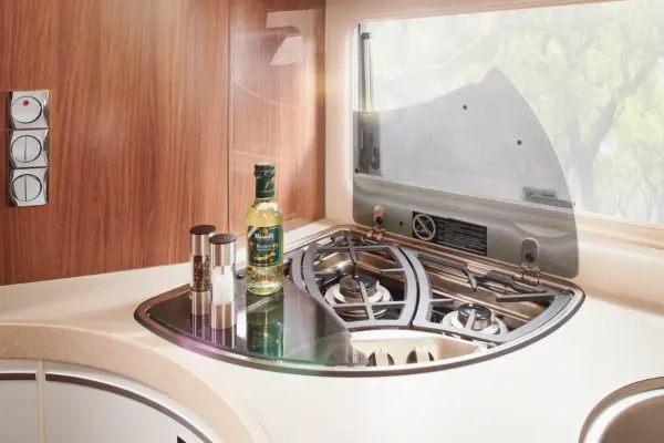 Eine kompakte Wohnmobilküche in der Carthago Chic E Line mit einem modernen Gasherd mit drei Brennern, einer hochgeklappten Glasabdeckung, Salz- und Pfefferstreuern und Olivenöl auf der Theke, alles beleuchtet vom natürlichen Licht des Fensters.