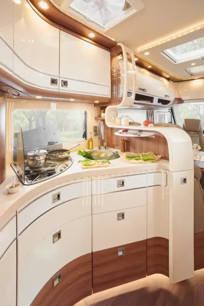 Moderne Wohnmobilküche in einem Carthago Chic S Plus mit glänzend weißen und hölzernen Schränken, einem Herd mit einem Topf, Gemüse und Lebensmitteln auf der Theke und hellem Tageslicht, das durch Oberlichter und Fenster einfällt.