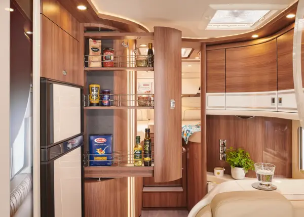 Eine moderne Wohnmobilküche im Carthago Chic S Plus verfügt über Holzschränke, eine offene, ausziehbare Speisekammer mit Lebensmitteln, einen Kühlschrank und eine kleine Arbeitsfläche mit einer Pflanze und einem Glas Wasser. Die warme Beleuchtung unterstreicht den organisierten Raum.