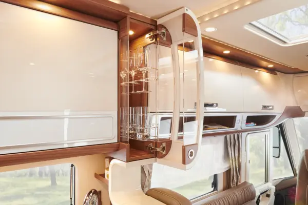 Ein offener Schrank in einem luxuriösen Carthago Chic S Plus Wohnmobil offenbart ordentlich arrangierte Gläser und Champagnerflöten. Die moderne Holz- und Weißverkleidung und die sanfte Beleuchtung schaffen eine elegante Atmosphäre.