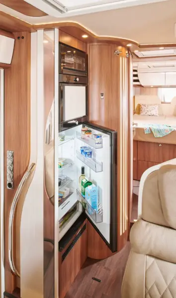 Ein modernes Carthago Chic S Plus Wohnmobil-Interieur mit einem offenen Kühlschrank, der mit Lebensmitteln und Getränken gefüllt ist, Holzschränken und einem Blick auf einen gemütlichen Schlafbereich mit Kissen und Decken im Hintergrund.