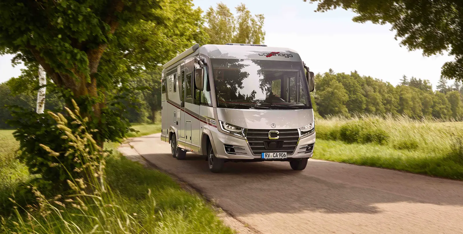 Ein silbernes Wohnmobil fährt an einem sonnigen Tag eine schmale Landstraße entlang, die von Bäumen und grünen Feldern gesäumt ist - perfekt für eine entspannte Probefahrt.