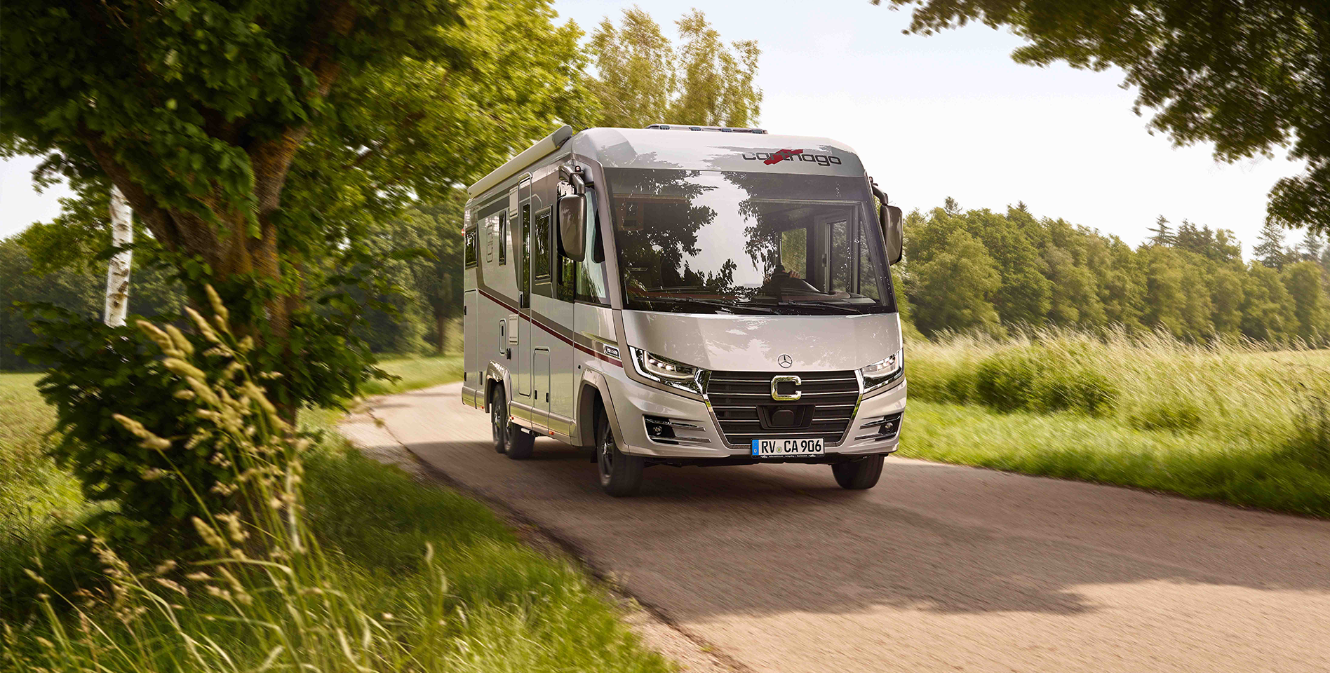 Ein silbernes Wohnmobil fährt an einem sonnigen Tag eine schmale Landstraße entlang, die von Bäumen und grünen Feldern gesäumt ist - perfekt für eine entspannte Probefahrt.