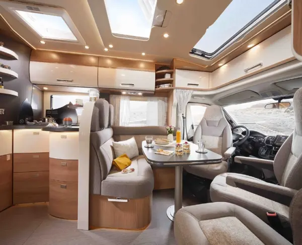 Modernes Wohnmobil-Interieur mit grauen und hölzernen Oberflächen, einem Esstisch mit Frühstück, gemütlichen Sitzgelegenheiten, Stauraum über Kopf, einem Dachfenster und großen Fenstern, die natürliches Licht hereinlassen. Der Fahrerbereich ist an der Vorderseite sichtbar.