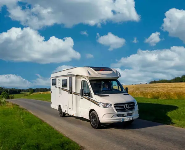 Ein weißes Mercedes-Benz Wohnmobil fährt auf einer schmalen Landstraße, umgeben von grünen Feldern und goldenem Gras unter einem strahlend blauen Himmel mit vereinzelten Wolken.